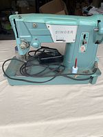 Full view of Singer teal sewing machine with foot pedal and cords on bed