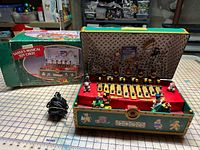 Music box open showing interior figures and xylophone mechanism, power adapter and product box visible