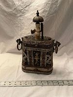 Front view of ornate mixed-metal vessel showing filigree panels, stone inlays, spout, bone finial and side ring handles with ruler for scale