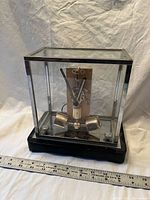 Front view of clear glass case clock with exposed movement and cylindrical pendulum on black base next to ruler for scale