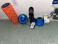 All items lined against wall on carpet: orange foam roller, blue exercise ball, massage gun, blue massage base, Up & Up tape