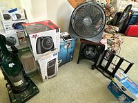 Group shot showing vacuum, boxed heaters, boxed pedestal fan, box fan, desk fan and CD rack