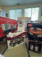 Stack of five boxed appliances: GE hot plate, Rival skillet, Parini pizza cooker, Parini rice cooker, Air Whirl Crisper lid