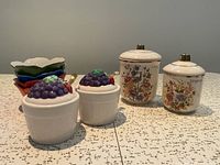 Group shot of all items: two floral canisters, two jam pots with grape lids and spoons, four coloured bowls
