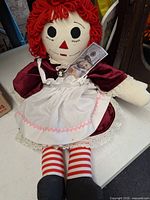 Full view of Raggedy Ann cloth doll wearing burgundy dress, white apron, striped legs, black shoes, replica $100 bill visible