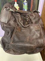 Front view of brown leather Rugby bucket backpack showing logo patch, front pocket, drawstring