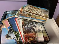 Open metal tin showing large unsorted stack of vintage color postcards with various scenic and architectural images