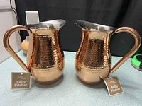 Front view of two hammered copper belly pitchers with tags