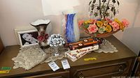 Overview of all items on dresser: art glass vase, ceramic vase, metal bouquet figurine, two doilies beneath, two boxes of taper candles