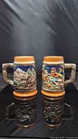 Front view of both ceramic steins showing raised-relief scenes and tan rims/bases