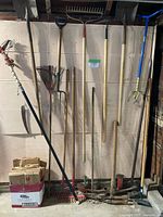 Overview of all long-handled tools lined against wall plus box of yard waste bags