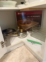 Contents inside cabinet: pizza stone box, two saucepans with lids, white casserole with lid, glass baking dishes