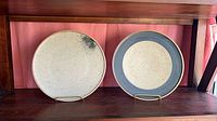Front view of one beige speckled plate and one blue-rim plate on stands