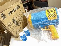 Box marked Rocket 697L with bubble graphics, bubble-wrapped blue and yellow bubble gun, two blue solution bottles