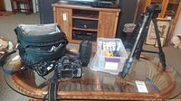 Wide shot of entire lot including camera with lens, extra lens, tripod, plastic bin with filters/accessories and camera bag on table