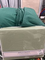 Boxed Hillcrest sheet set in light green stripe packaging beside stacked dark green loose linens