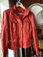 Front view of red leather jacket on hanger showing pockets, buttons and epaulettes