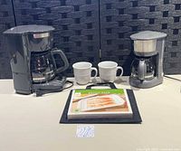 Both coffee makers, two mugs, bacon rack package atop black pad
