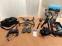 Overhead view of entire lot including DJI drone, remote, two batteries, Nikon case, Bushnell binoculars, Bower kit box and components, tabletop tripod, and selfie stick