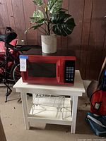 Front view of all items: red microwave on white stand with wire rack below and faux plant on top