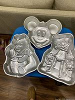 Three aluminum character cake pans displayed: Mickey Mouse, Winnie the Pooh, girl with doll