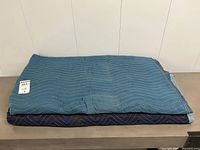 Two folded moving blankets stacked, teal on top, navy beneath