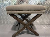 Front view of upholstered ottoman bench showing linen seat, nailhead trim, X-base legs