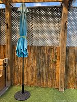 Full view of closed aqua/teal umbrella on black pole in round base against lattice backdrop