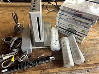 Console, remotes, cables, sensor bar, stand, and stack of game cases displayed on workbench