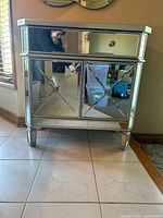 Front view of mirrored accent cabinet showing drawer, cabinet door, mirrored X-motif, silver frame