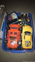 Bin filled with small die-cast cars, two large plastic cars, handheld game visible