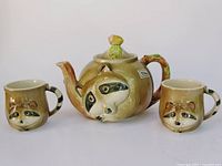 Front view of teapot and two mugs showing raccoon faces