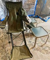 Green folding camping chair with footrest, carry bag of second chair, two glass top tables visible