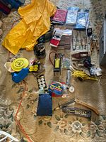 Array of camping items laid out including ponchos, rods, saw, pot, pump, tackle box, rope, tarps, candles and disc