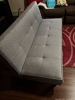 Futon shown in sofa position, light gray fabric, armless, button tufting, visible fraying at lower corner