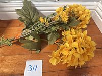 Three faux stems with yellow flower heads and green leaves laid on hardwood floor, paper lot number visible