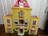 Front view of closed dollhouse showing yellow facade, pink roof, windows, balcony