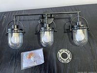 overall view of black metal three-light vanity fixture, glass shades, LED bulbs, mounting plate and hardware pack on surface