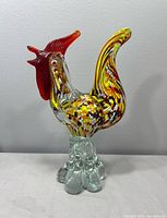 Full view left side of multicolored glass rooster on clear base
