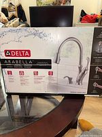 Front of sealed retail box showing Delta Arabella kitchen faucet model and features