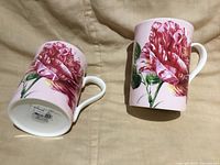 Pair of rose-pattern ceramic mugs on fabric background, one upright and one on side showing base label