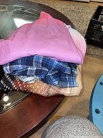 Stack of four folded women’s garments: pink T-shirt, pastel top, blue plaid shirt, brown polka-dot blouse