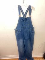 Front view of medium blue denim overalls on hanger showing bib, pocket, straps and legs