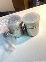 Two stainless steel shakers side by side, one partially wrapped in plastic, showing lids