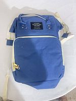 Front view of royal blue Traveling Share backpack showing brand label, front zip pocket, white trim, beige straps