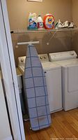 Laundry area scene showing three steam irons on shelf and full-size ironing board leaning against washer