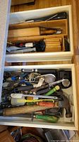 Open drawers containing assorted kitchen utensils and small hand tools, including hammer, screwdrivers, pliers, wrench, can opener, peeler, scoops, digital timer, rolling pin and knife blocks