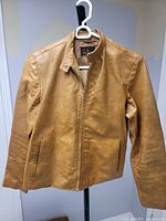 Front view of tan leather jacket on hanger showing zipper closure, pockets, collar