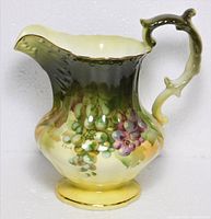 Front view showing painted grape design, scalloped rim and ornate handle