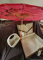 Suitcase open showing interior straps and keys, parasol set above, wicker basket alongside